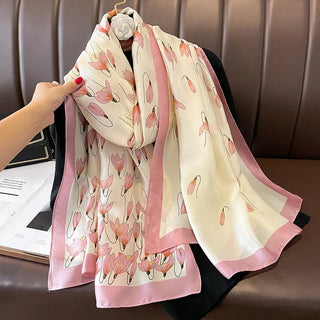 A long satin silk scarf with a floral pattern in pink and white colors, draped over a chair with a person's hand adjusting its position.